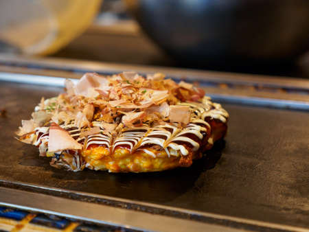 Closeup detail of a freshly cooked Okonomiyaki topped with Katsuobushi fish flakes, sauce, and mayonnaise, on a Teppan grill. Shallow focus. Dotonbori, Osaka, Japan. Travel and cuisine.の写真素材