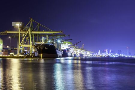 Container cargo ship with ports crane bridge in harbor and refinery industrial at night logistics...