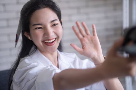 beautiful young asian woman smiling while selfie in front of camera against white brick wallの写真素材