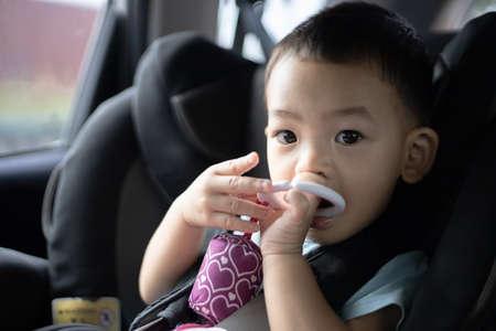 1 Year Old Adorable, an Asian Toddler Boy, Sitting in a Car Seat on a Car While Traveling Looking at the camera. Family Trips and Baby Care and Safety Concept. Front View Closeup Blurry Background.の写真素材