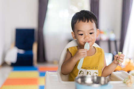 An Adorable toddler Asian baby boy (1-year-old) sitting on a high feeding chair eating homemade food using hands and spoon at home. Children's closeup portrait of activity and baby lifestyle concept.の写真素材