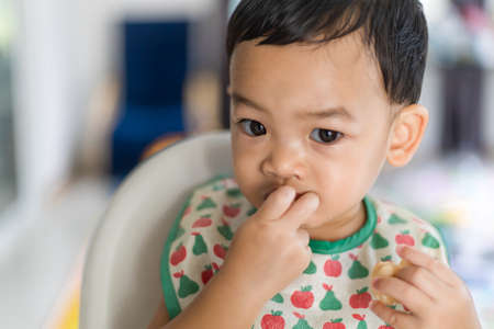 An Adorable toddler Asian baby boy (1-year-old) sitting on high feeding chair eating homemade food and learning to use hands at home. Children's closeup portrait of activity and baby lifestyle conceptの写真素材