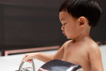 Adorable Asian kid boy (Toddler age 1-year-old) take a shower in the white bathtub by himself,...