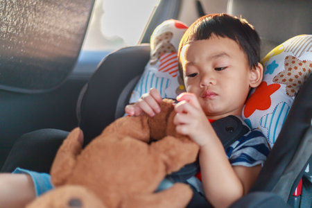 Adorable Asian kid boy (Toddler age 1-year-old) Protection Sitting in the Car Seat with Safety Belt Locked and Holding Bear Doll. Journey Baby Careful Concept. Portrait CloseUp and Blurry Background.の写真素材