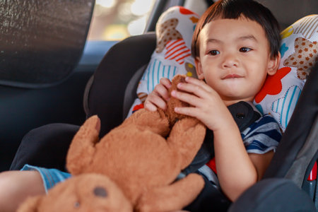 Adorable Asian kid boy (Toddler age 1-year-old) Protection Sitting in the Car Seat with Safety Belt Locked and Holding Bear Doll. Journey Baby Careful Concept. Portrait CloseUp and Blurry Background.の写真素材