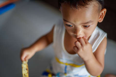 An Adorable toddler Asian baby boy (1-year-old) sitting and self-feeding homemade pancake food and learning to use hands at home. Children's closeup portrait of activity and baby lifestyle concept.の写真素材