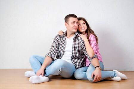 Beautiful barefoot young couple in casual jeans sitting leaning against a white wall on the wooden floor in the living room smiling at the cameraの写真素材