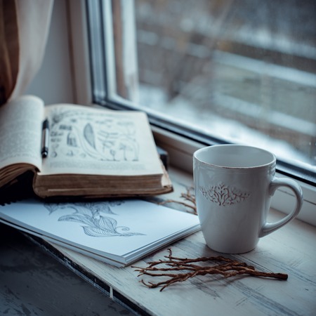Cozy winter still life: cup of hot coffee and opened book on vintage windowsill against snow landscape from outside.の写真素材