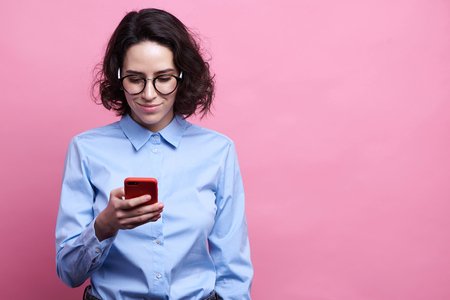 Glad attractive female with dark skin and curly black hair standing indoors with her smartphone chatting with friends. Happy cute woman in casual clothes recieving messages from her boyfriendの写真素材