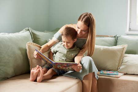 Young mother, read a book to her child, boy in the living room of their home, rays of sun going through the windowの写真素材