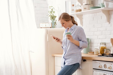 Young woman using smartphone leaning at kitchen table with coffee mug and organizer in a modern home. Smiling woman reading phone message. Brunette happy girl typing a text messageの写真素材