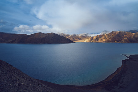 A panoramic view of Pangong Lake during golden hour in Ladakh, India, with snow-capped mountains illuminated by sunlight and crystal-clear water creating a peaceful and majestic Himalayan landscapeの写真素材