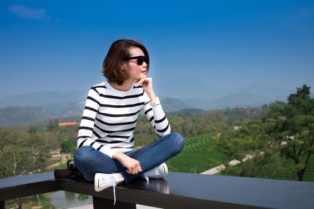 Beautiful Asian woman sitting outdoors on black bench at green tea farm against blue skyの写真素材