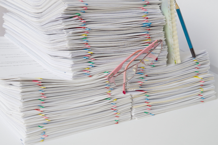 Pink spectacles in front of pile of overload paperwork and notebook with blue pencil on white table.の写真素材