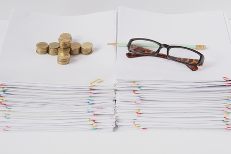 Spectacles and pencil with pile of gold coins place on pile of overload paper and reports with colorful paper clip on white table.の写真素材