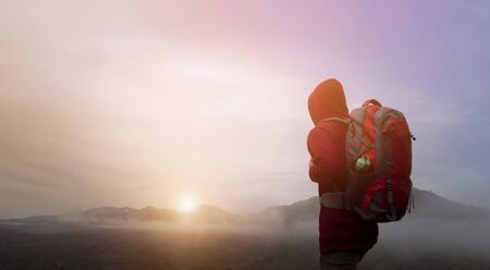 Back view of man traveler with backpack walking on path and heading to foggy mountain and sunrise in...