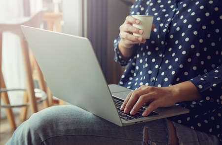 Young woman relaxing and drinking cup of hot coffee or tea using laptop computer in the cafe. Cozy...
