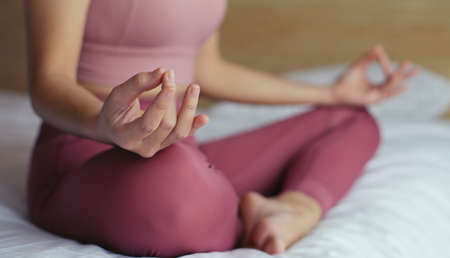 Close-up hand of young woman sitting in lotus pose on bed at home. Morning workout in bedroom.