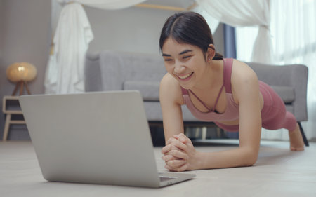 Young strong woman is training at home, standing in plank position, workout with online trainer...