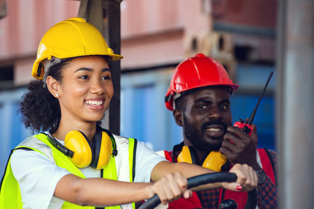 Two African American male and female worker in uniform and helmet driving and operating on diesel container forklift truck at commercial dock site.の写真素材
