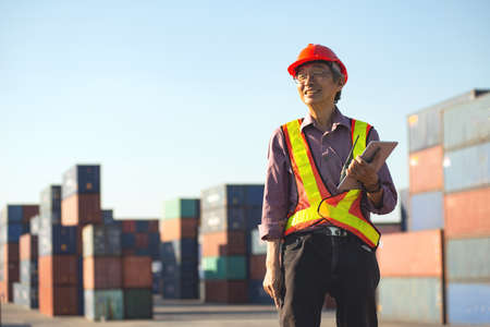 A senior elderly Asian worker engineer wearing safety vest and helmet standing and holding digital tablet at shipping cargo containers yard.の写真素材