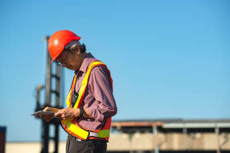 A senior elderly Asian worker engineer wearing safety vest and helmet standing and holding digital...