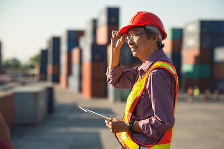 A senior elderly Asian worker engineer wearing safety vest and helmet standing and holding digital tablet at shipping cargo containers yard.の写真素材