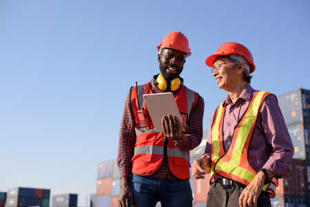 Happy African American and senior asian male worker using tablet during video call in a logistic...