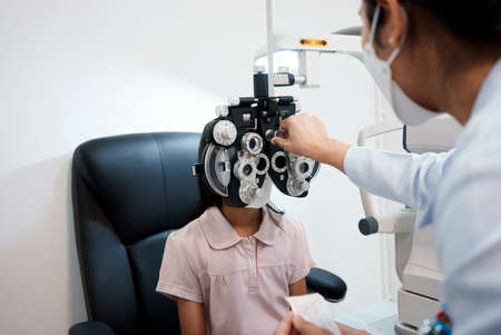 Ophthalmologist examining the eyes of an Asian girl in a clinic. They wear protective face masks.の写真素材