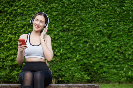 Young woman in sportswear listening music with headphones on  park. She sitting on a bench while resting and using smartphone.の写真素材
