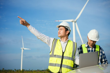 Two asian engineers discussing and checking turbines on wind turbine farm. Renewable energy technology and sustainability.の写真素材