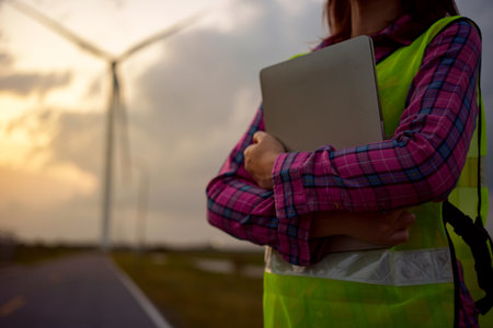 Asian woman working with laptop at renewable energy farm. Female inspector controlling functioning of wind turbines outdoors.の写真素材