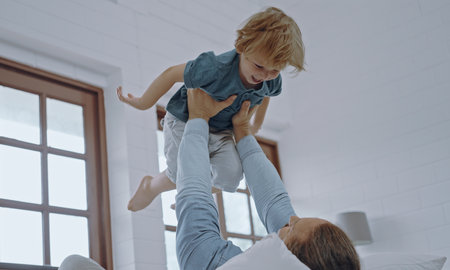 Happy kid boy flying in father's arms in bedroom.father holding raising cute little son playing airplane bonding having fun in bed.の写真素材