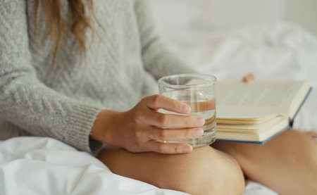 Young asian woman drinking water while reading a book sitting on the bed in the morning.の写真素材