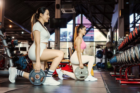 Two young asian woman doing exercises with heavy weights plates in gym.