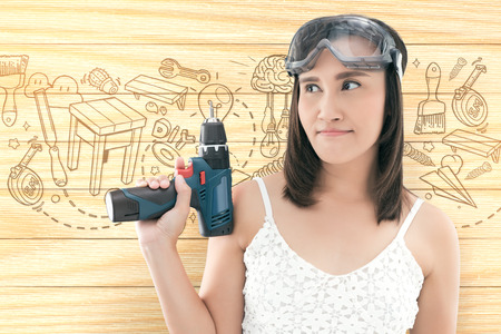 Asian woman dressed in white top holding a cordless electric drill on wood background