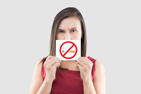Asian woman in red blouse is holding no smoking sign on the white paper against gray background. Picture of the woman with a smoking restriction sign. Warning about the harm of tobacco use at the public place.の写真素材