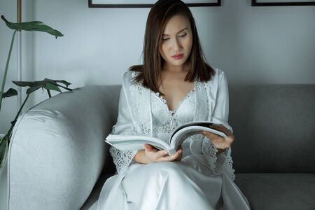 Asian women wear a satin nightgown and white lace robes reading the magazine article on a gray sofa at night before going to sleep.の写真素材