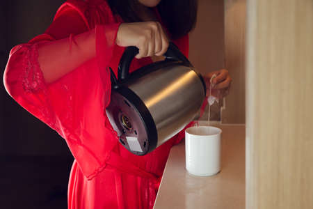 The woman wore red satin long robe making tea in the kitchen at night. Asian girl pour hot water into a white cupの写真素材