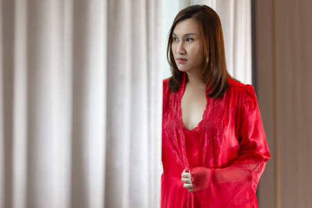 Asia woman in a red satin nightgown standing side window and looking at skyscrapers out the window in the morning.の写真素材