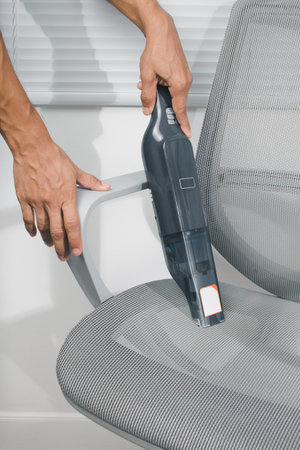 The man is using a cordless car vacuum cleaner to clean the gray chairs in the office.の写真素材
