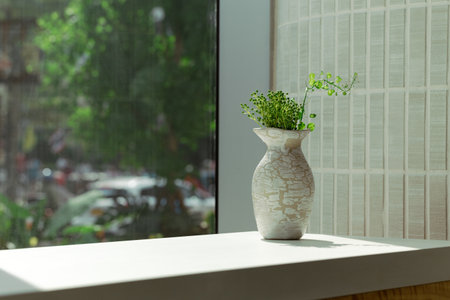 A beautifully crafted vase with intricate patterns holds green plants on a sunlit windowsill; the outside view through the window adds a touch of nature to the serene indoor setting.の写真素材