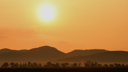 Picture of a sun setting behind a dense forest area followed by mountains.の写真素材