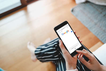 Bangkok, Thailand - MAY 9, 2020: A woman sit on the bad within bedroom near glass door for using iphone 7 plus with Google search engine on web page.のeditorial素材