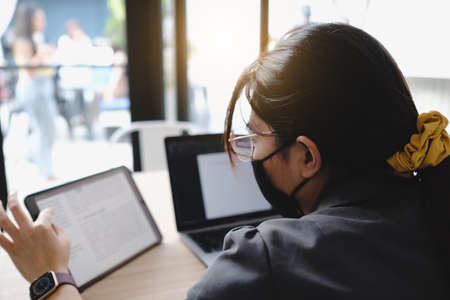 Closeup image of a beautiful Asian business woman wear mark holding , using and looking at smart phone with laptop and tablet pc on wooden table in modern cafeの写真素材