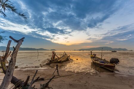 Fishing boat at the beach (long-tail boats) and boat carcass in low water and the beautiful sunrise at Phuket Island, Thailand.の写真素材