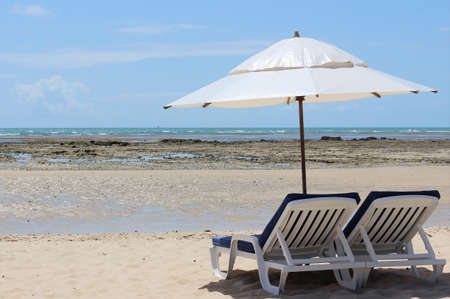 Beach with a pair of white plastic loungers, blue cushions and white parasol on the sand and with the sea and blue sky in the background. Morro de Sao Paulo, Cairu, Bahia, Brazil.の写真素材