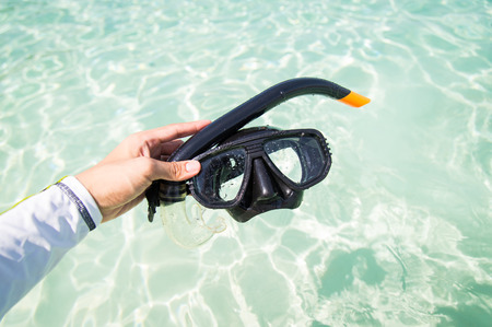 Hand holding snorkel goggles against beach and sea surface.の写真素材