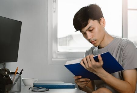 Young handsome asian man reading book at work desk in free time from working at home, Knowledge and...