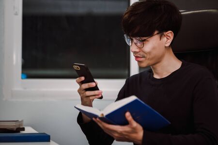 Young handsome asian man playing smartphone and reading book at work desk late at night, Knowledge...